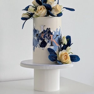 Puede incluir: Un pastel blanco con un diseño abstracto azul y plateado y las iniciales "J & K" en la parte delantera. El pastel está cubierto con rosas blancas y hojas azules y está sentado en un soporte para pastel blanco.