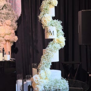 Puede incluir: Un pastel de bodas blanco de cuatro pisos decorado con aliento de bebé blanco y rosas blancas. El nivel superior tiene las iniciales "J & M" en letras doradas.