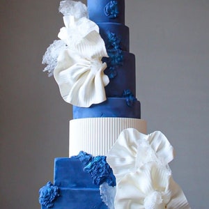 Könnte beinhalten: Eine hohe, blaue Hochzeitstorte mit weißen Fondantblumen und filigranen Details. Die Torte ist in einem blauen und weißen Farbschema dekoriert.