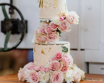 Puede incluir: Un pastel de boda de cuatro pisos decorado con rosas rosadas y flores blancas. El pastel es blanco con detalles dorados y las iniciales "M & J" están en el tercer piso.