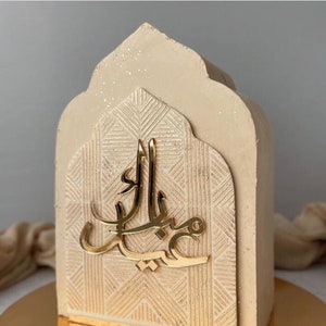 May include: A beige concrete cake decorated with a gold geometric pattern and a gold Arabic inscription "Eid Mubarak".