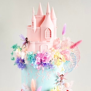 Könnte beinhalten: Eine pastellrosa und blaue Feentorte mit einem rosa Schloss oben, umgeben von pastellfarbenen Blumen und Feenfiguren. Die Torte ist mit einer Holztür verziert, auf der "Willkommen im Feenland" steht.