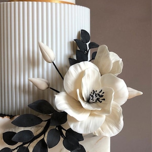 Może przedstawiać: Biały tort ślubny ozdobiony czarnymi liśćmi i białymi kwiatami. Tort ma złotą wstążkę i marmurowy wzór.