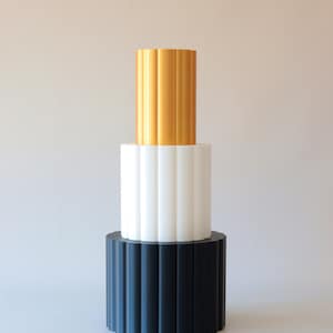 Könnte beinhalten: Drei übereinander gestapelte zylindrische Sockel mit einem geriffelten Design. Der obere Sockel ist golden, der mittlere Sockel ist weiß und der untere Sockel ist schwarz.