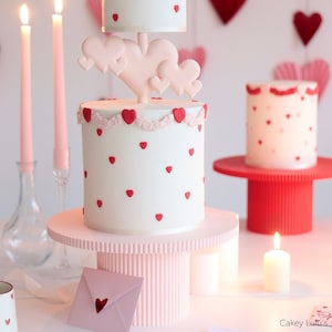 Könnte beinhalten: Eine zweistöckige Hochzeitstorte, verziert mit rosa und roten Herzen. Die obere Etage ist weiß mit roten Herzen und einem rosa-weißen Rand. Die untere Etage ist weiß mit roten Herzen und einem rosa-weißen Rand. Die Torte steht auf einem rosa Tortenständer mit einem rosa-weißen gestreiften Sockel. Auf dem Tisch stehen Kerzen und ein rosa Umschlag mit einem roten Herz darauf.
