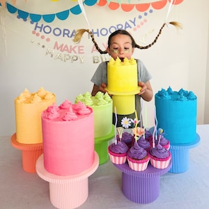 Könnte beinhalten: Eine Regenbogen-Geburtstagstorten-Anzeige mit einer gelben Torte, einer rosa Torte, einer grünen Torte und einer blauen Torte. Jede Torte steht auf einem mehrstufigen Ständer. Es gibt auch Cupcakes mit lila Frosting und bunten Streuseln. Der Text auf dem Banner lautet "Hooray Make a Happy Birthday".