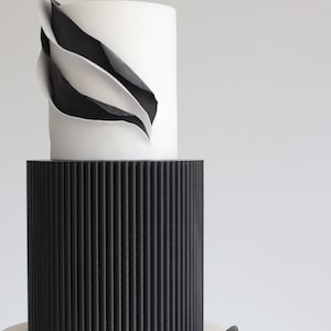Könnte beinhalten: Eine dreistöckige Hochzeitstorte mit schwarzem und weißem Fondant. Die oberste Schicht hat eine weiße Fondantbasis mit einem schwarzen und weißen abstrakten Design. Die mittlere Schicht ist schwarzer Fondant mit vertikalen Linien. Die unterste Schicht ist weißer Fondant.
