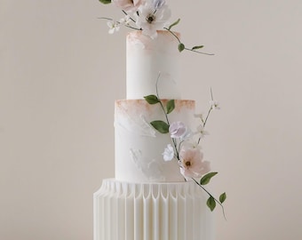 May include: A three-tiered white cake with floral decorations. The cake has a textured design on the bottom tier and a gradient of peach and white on the top tiers. Delicate flowers and greenery cascade down the sides.