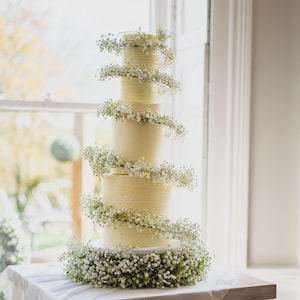 Könnte beinhalten: Mehrstöckige Hochzeitstorte mit weißer Glasur und zarten floralen Akzenten. Jede Schicht ist mit einer Spirale aus kleinen weißen Blüten geschmückt, die ein elegantes und festliches Design schaffen. Die Torte steht auf einem Tisch mit weißem Tuch.