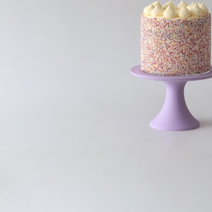 Puede incluir: Un soporte para pastel de un solo nivel con una base morada y una parte superior redonda. Un pequeño pastel redondo con glaseado blanco y chispas de colores está en la parte superior del soporte.