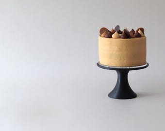 May include: A single-tiered cake with a light brown frosting and chocolate and caramel decorations on top. The cake is on a black cake stand.