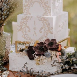 May include: A four-tiered white wedding cake with gold accents and floral decorations. The cake is decorated with a delicate floral pattern and the initials "I & A" on the third tier. The cake is displayed on a gold stand and surrounded by dried flowers.