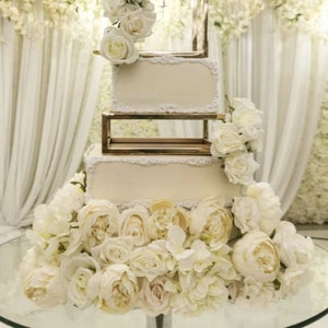 May include: A white square wedding cake with gold accents and white flowers. The cake is on a glass cake stand and is surrounded by white flowers.