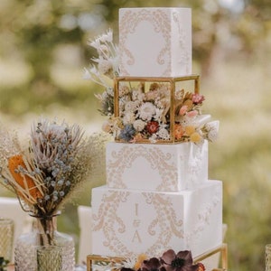 Peut inclure: Un gâteau de mariage à quatre étages avec un glaçage blanc et des accents dorés. Le gâteau est décoré de fleurs séchées et de verdure. Le niveau supérieur a une cage dorée avec des fleurs séchées à l'intérieur. Le gâteau est sur un support doré. Le gâteau est décoré des initiales "I & A" sur le troisième étage.