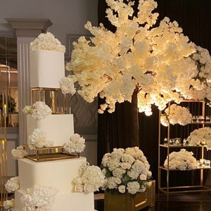 Peut inclure: Un gâteau de mariage blanc à quatre étages décoré de fleurs blanches et d'accents dorés. Le gâteau est présenté sur un support en acrylique transparent avec des accents dorés. Le gâteau est devant un fond noir avec deux arbres à fleurs blanches et une étagère en métal doré avec des fleurs blanches.
