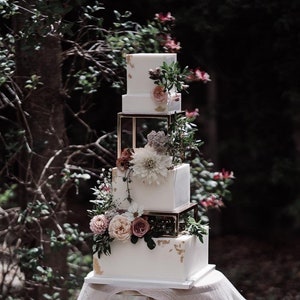 Peut inclure: Un gâteau de mariage à quatre étages décoré de glaçage blanc et d'accents dorés. Le gâteau est orné de fleurs roses et blanches et de verdure, et est présenté sur un support à gâteau blanc.
