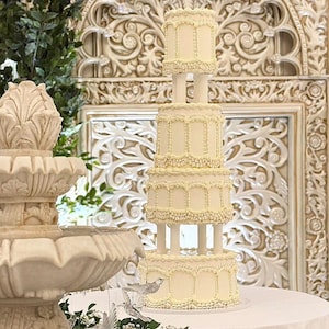 Puede incluir: Una tarta de boda crema de cinco pisos con intrincados detalles de glaseado, exhibida sobre una mesa redonda cubierta con un mantel blanco. Una fuente decorativa y paneles de pared tallados adornan el fondo, con acentos verdes.