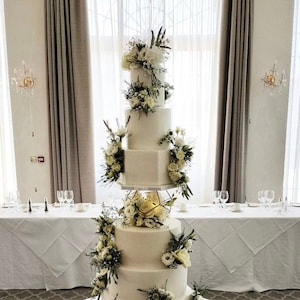 May include: A white wedding cake with geometric tiers decorated with white flowers and greenery. The cake is on a gold stand and is surrounded by white tablecloths.