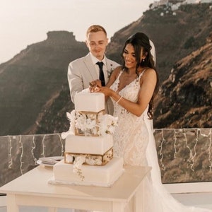Peut inclure: Une mariée et un marié coupant un gâteau de mariage blanc avec des accents dorés. Le gâteau est décoré de fleurs blanches et se trouve sur une table blanche. Le couple est debout devant un paysage pittoresque d'un village blanc et de montagnes.