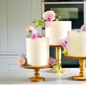 May include: Three white cakes decorated with pink and purple flowers, displayed on wooden cake stands of varying heights.