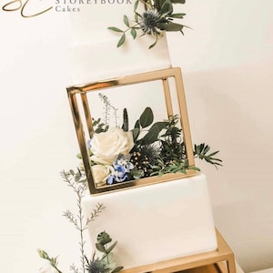 Peut inclure: Un gâteau de mariage blanc avec des étages géométriques dorés décorés de roses blanches, de fleurs bleues et de verdure.