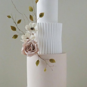 Könnte beinhalten: Eine dreistöckige Hochzeitstorte mit einer rosafarbenen unteren Etage, einer weißen, strukturierten mittleren Etage und einer weißen oberen Etage. Die Torte ist mit einem zarten Blumenarrangement aus weißen und rosafarbenen Blüten und braunen Zweigen verziert.