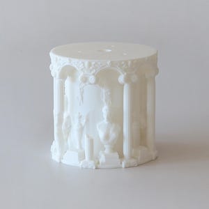 Może przedstawiać: Biały, cylindryczny świecznik o klasycznym architektonicznym designie. Świecznik posiada kolumny, łuki i rzeźbione figury, tworząc estetykę miniaturowej świątyni. Na górze znajdują się trzy małe otwory. Całkowity design jest szczegółowy i ozdobny.