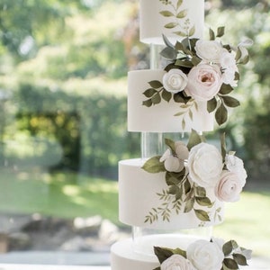 May include: A white wedding cake with four tiers, decorated with delicate sugar flowers and green foliage. The cake is displayed on clear acrylic stands.