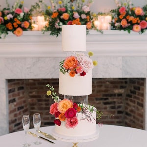 May include: A three-tiered white wedding cake decorated with pink, orange, and red flowers. The cake is on a gold geometric stand and sits on a white tablecloth.