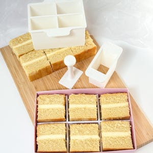 Könnte beinhalten: Ein weißer quadratischer Kuchenausstecher aus Kunststoff mit vier Fächern, ein rechteckiger Ausstecher und ein weißer Stempel. Sechs quadratische Kuchenstücke mit Cremefüllung liegen in einer rosa Schachtel, weitere Stücke auf einem Holzbrett.