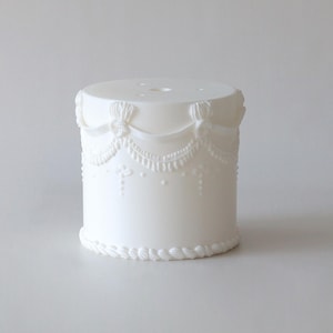 Könnte beinhalten: Ein weißer, zylindrischer Tortenständer mit aufwendigen, erhabenen Verzierungen. Das Design umfasst Schleifen, Girlanden und einen seilartigen Rand an der Basis. Der Ständer besteht aus einem festen, undurchsichtigen Material.