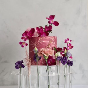 May include: A clear acrylic cake stand with a pink and white cake decorated with gold accents. The cake stand has multiple small clear tubes for holding flowers. The cake is surrounded by pink, purple, and white flowers.