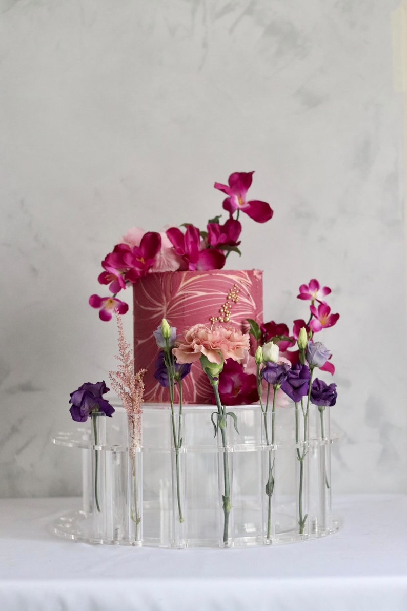May include: A clear acrylic cake stand with a pink and white cake decorated with gold accents. The cake stand has multiple small clear tubes for holding flowers. The cake is surrounded by pink, purple, and white flowers.