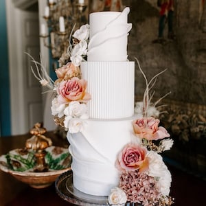 Könnte beinhalten: Eine dreistöckige weiße Hochzeitstorte mit Blumendekoration. Der Kuchen hat ein strukturiertes Design und ist mit rosa und weißen Rosen und anderen Blumen verziert. Der Kuchen steht auf einer transparenten Tortenplatte aus Glas.