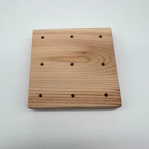 May include: A light-colored, rectangular wooden board with nine evenly spaced holes. The wood grain is visible, and the board has a slightly angled edge on one side. The board is likely for a craft or woodworking project.