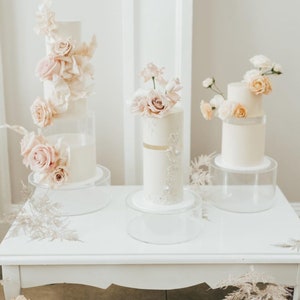 May include: Three white wedding cakes decorated with pink and peach flowers on clear acrylic cake stands. The cakes are on a white table with a white background.