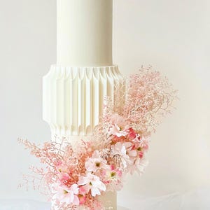 Könnte beinhalten: Eine zweistöckige weiße Hochzeitstorte mit einem geometrischen Design auf der unteren Etage. Die Torte ist mit rosa Blumen und Grün verziert.