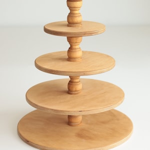 May include: A four-tier wooden cake stand with round tiers and turned wooden posts. The stand is made of natural wood and has a light brown finish.