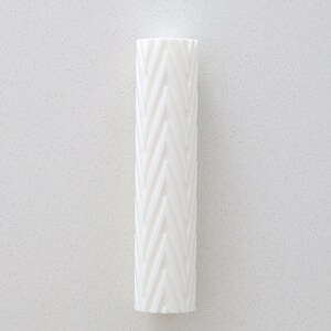 Könnte beinhalten: Eine hohe, weiße, zylindrische Vase mit einem strukturierten Fischgrätmuster. Die Vase besteht aus einem festen, undurchsichtigen Material und steht vertikal vor einem weißen Hintergrund. Das Design zeigt eine Reihe abgewinkelter Linien, die eine sich wiederholende V-Form bilden.