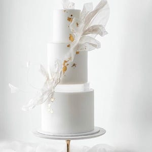 Peut inclure: Un gâteau de mariage blanc à trois étages avec des accents de feuilles d'or et un arrangement de fleurs en papier blanc sur le dessus. Le gâteau est sur un support doré.