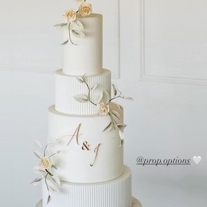 Könnte beinhalten: Eine mehrstöckige weiße Hochzeitstorte mit Blumen- und Blattdekorationen. Die Torte hat eine glatte Oberfläche mit gerippten Details auf einigen Etagen. Goldene Buchstaben "A&J" befinden sich auf der dritten Etage. Die Torte steht auf einem Marmorständer.
