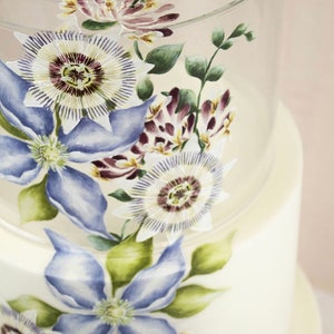 May include: A white wedding cake decorated with hand-painted blue and white flowers. The cake is displayed on a clear acrylic cake stand.