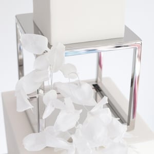 May include: A three-tiered cake stand with a white finish. The top tier is square, the middle tier is square, and the bottom tier is rectangular. The cake stand is made of metal with a silver finish. White silk flowers are draped over the cake stand.