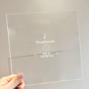 Może przedstawiać: Przezroczysty kwadratowy arkusz akrylowy z nadrukowanym tekstem "PropSecure" i "PROP OPTIONS". Wewnątrz znajduje się okrąg z liczbą "8".