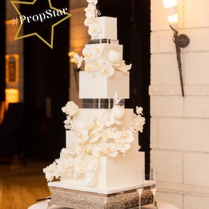 Może przedstawiać: Biały kwadratowy tort ślubny ozdobiony białymi orchideami. Tort stoi na srebrnym stojaku i ma wzór w biało-czarną kratę. Tort stoi na białym stole z dwoma kieliszkami do szampana. Tekst "With Love By Esther James" znajduje się na dole obrazu.