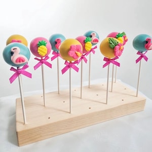 May include: A wooden cake pop stand with 12 cake pops decorated with tropical themes. The cake pops are decorated with pink flamingos, pineapples, and flowers. The cake pops are on white sticks with pink bows.