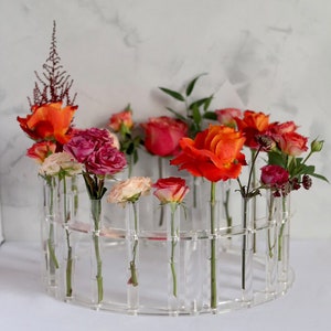 May include: A clear acrylic flower vase with a circular design holds a variety of roses in shades of pink, orange, and red. The vase has multiple small tubes for each flower stem.