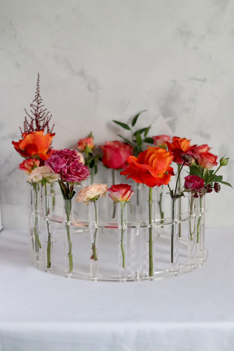 May include: A clear acrylic flower vase with a circular design holds a variety of roses in shades of pink, orange, and red. The vase has multiple small tubes for each flower stem.