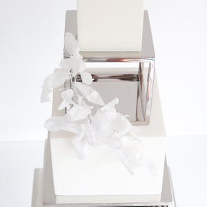 May include: A white cake with four tiers, each tier is decorated with a silver square frame. The cake is decorated with a white floral garland.