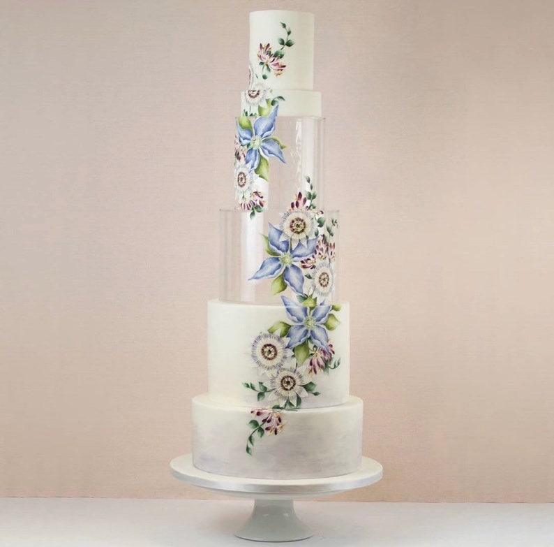 May include: A white wedding cake with clear acrylic tiers decorated with hand-painted floral designs in shades of blue, white, and green.